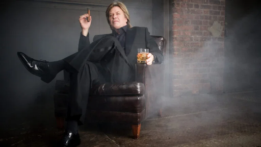 Ron White seated with a cigar and whiskey in a smoky room